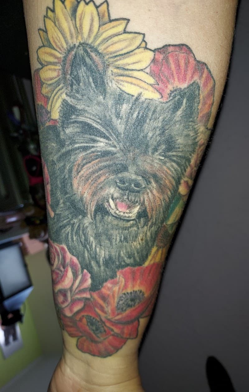 Illustratieve tattoo van een zwarte terriër met zonnebloem en klaprozen.