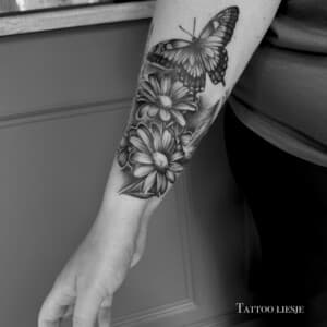 Zwart-wit tattoo van een vlinder en madeliefjes op de onderarm.