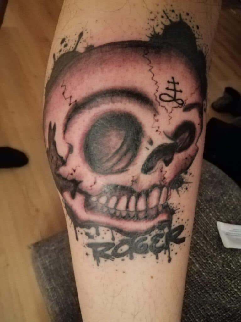 Schedel tattoo met 'Roger' tekst en symbool op onderbeen.