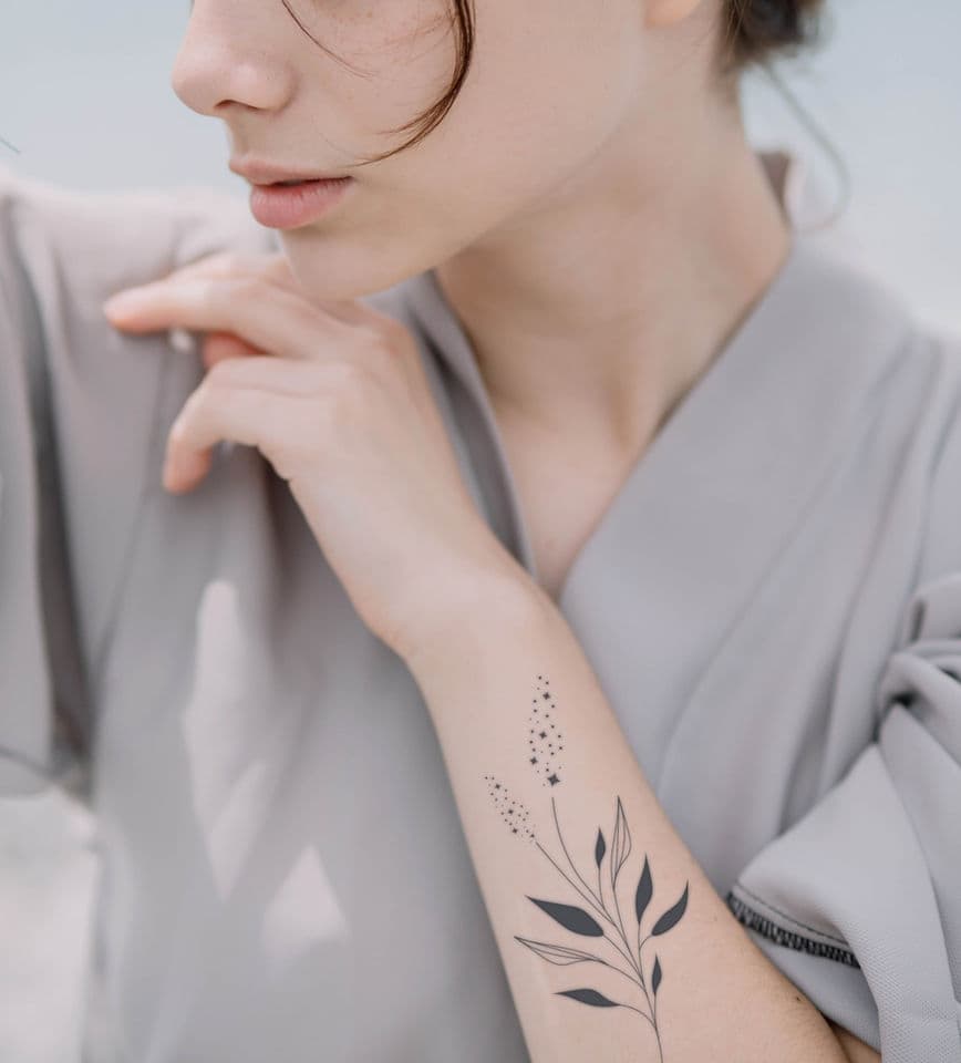 Fineline tattoo van een tak met bladeren en sterretjes op de onderarm.