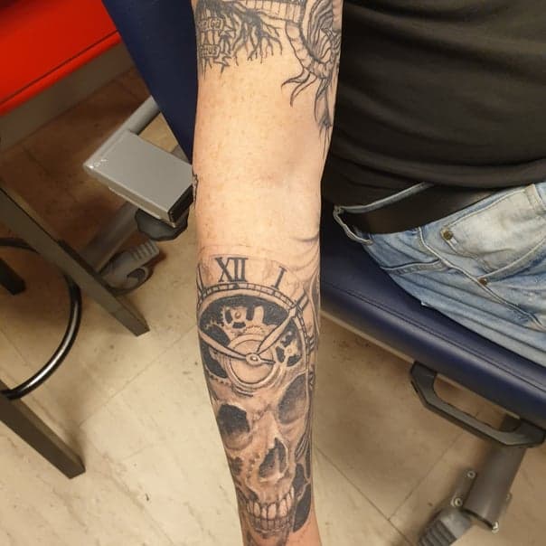 Zwart-wit tattoo van een schedel met een klok en tandwielen op de onderarm.