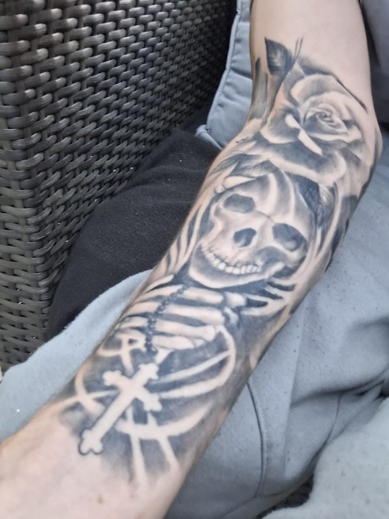 Zwart-wit tattoo van schedel, roos en rozenkrans op onderarm.