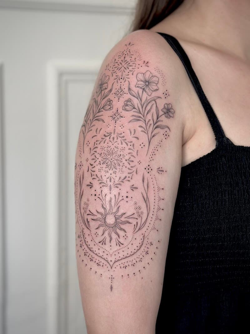 Fineline en dotwork tattoo met bloemen, mandala en zon op de bovenarm.