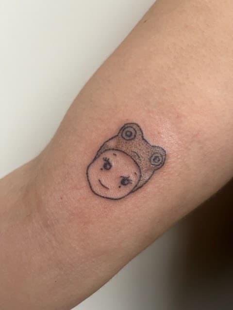 Kleine fineline tattoo van een gezichtje met een kikkerhoed op de arm.