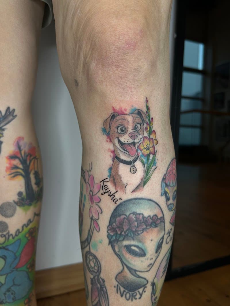 Kleurrijke cartoon hond 'Keysha' en alien 'Ivory' tattoos op onderbeen.