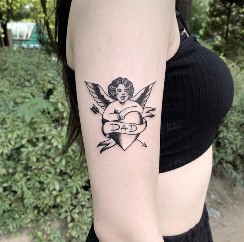 Blackwork tattoo van een cherubijn met een hart en 'DAD' banner op de bovenarm.
