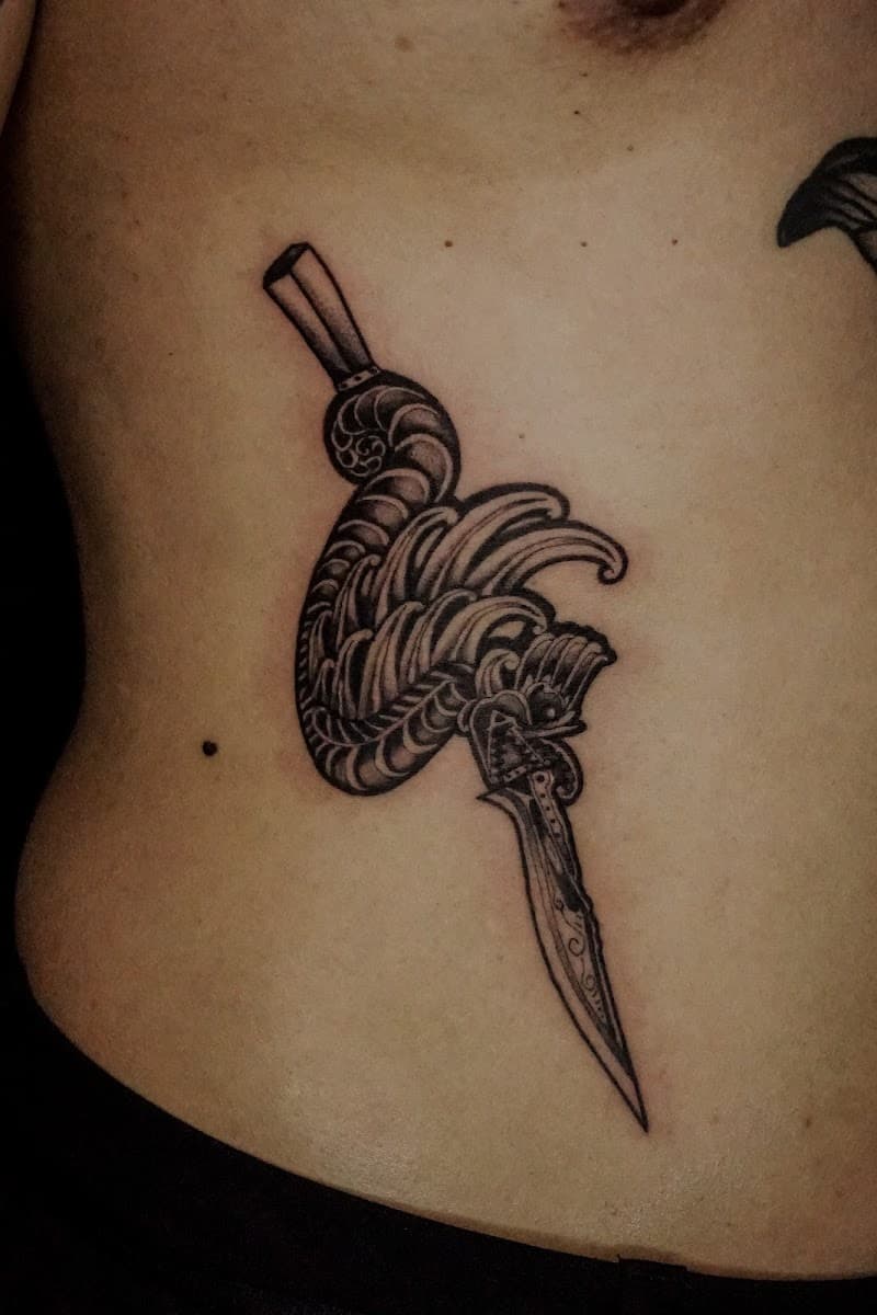 Zwart-wit tattoo van een dolk met een gedetailleerd, golvend handvat op de ribben.