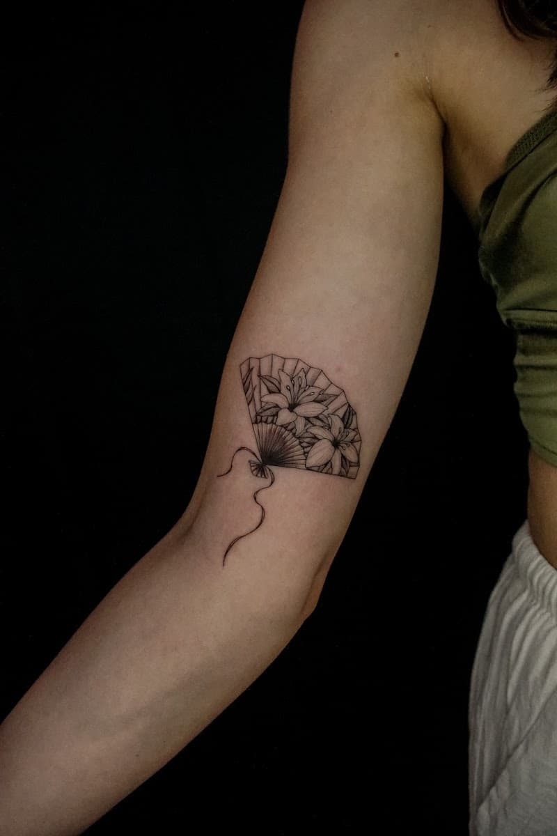 Fineline tattoo van een waaier met lelies op de bovenarm.