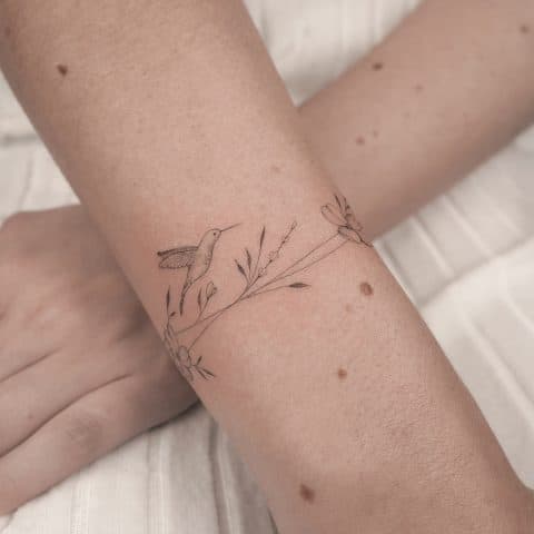 Fineline tattoo van een kolibrie met bloemen en takjes rond de pols.