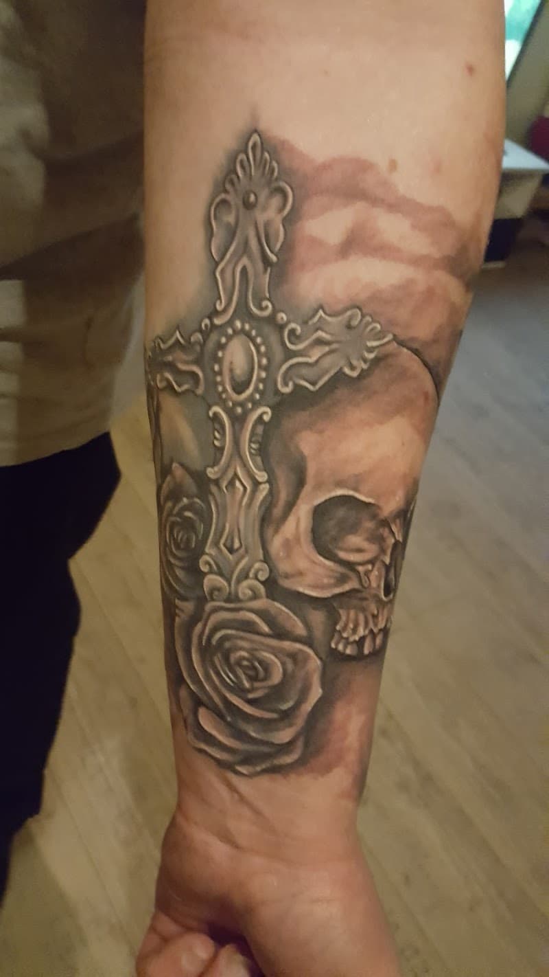 Zwart-wit tattoo van een gedetailleerd kruis, schedel en rozen op de onderarm.