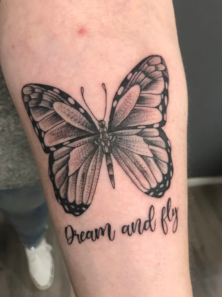 Zwart-wit vlinder tattoo met de tekst "Dream and fly" eronder.