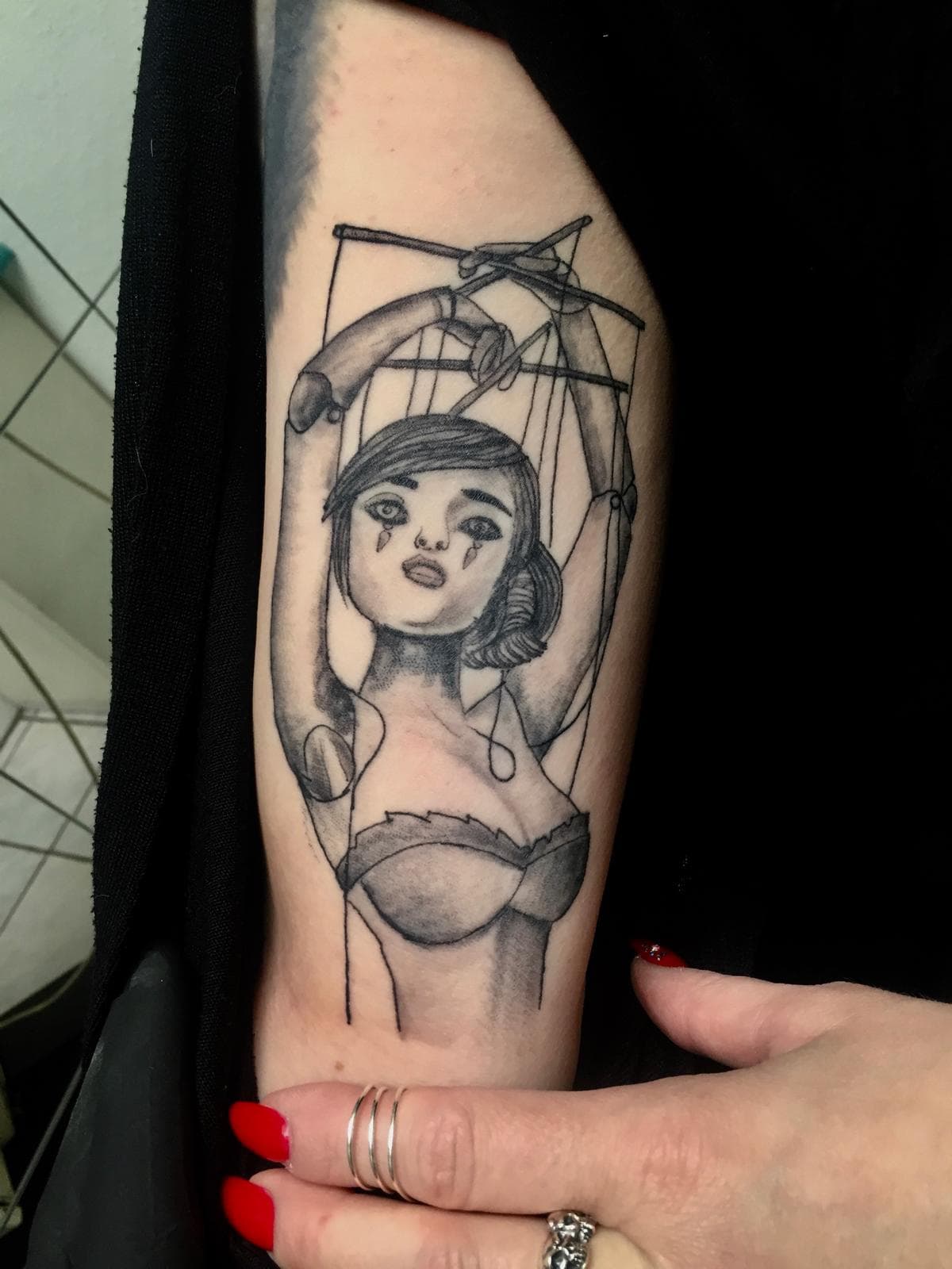 Zwart-wit tattoo van een huilende marionetvrouw aan touwtjes op de bovenarm.