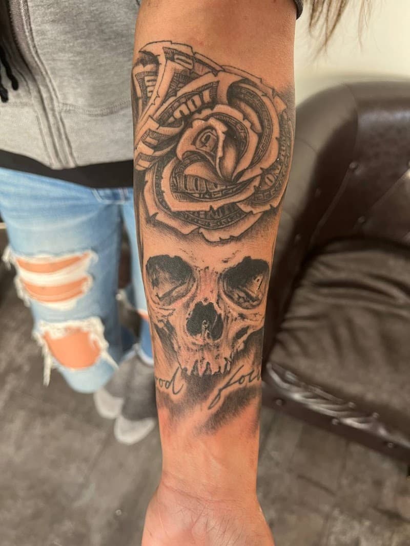 Zwart-grijze tattoo van een schedel met een roos van geld op de onderarm.