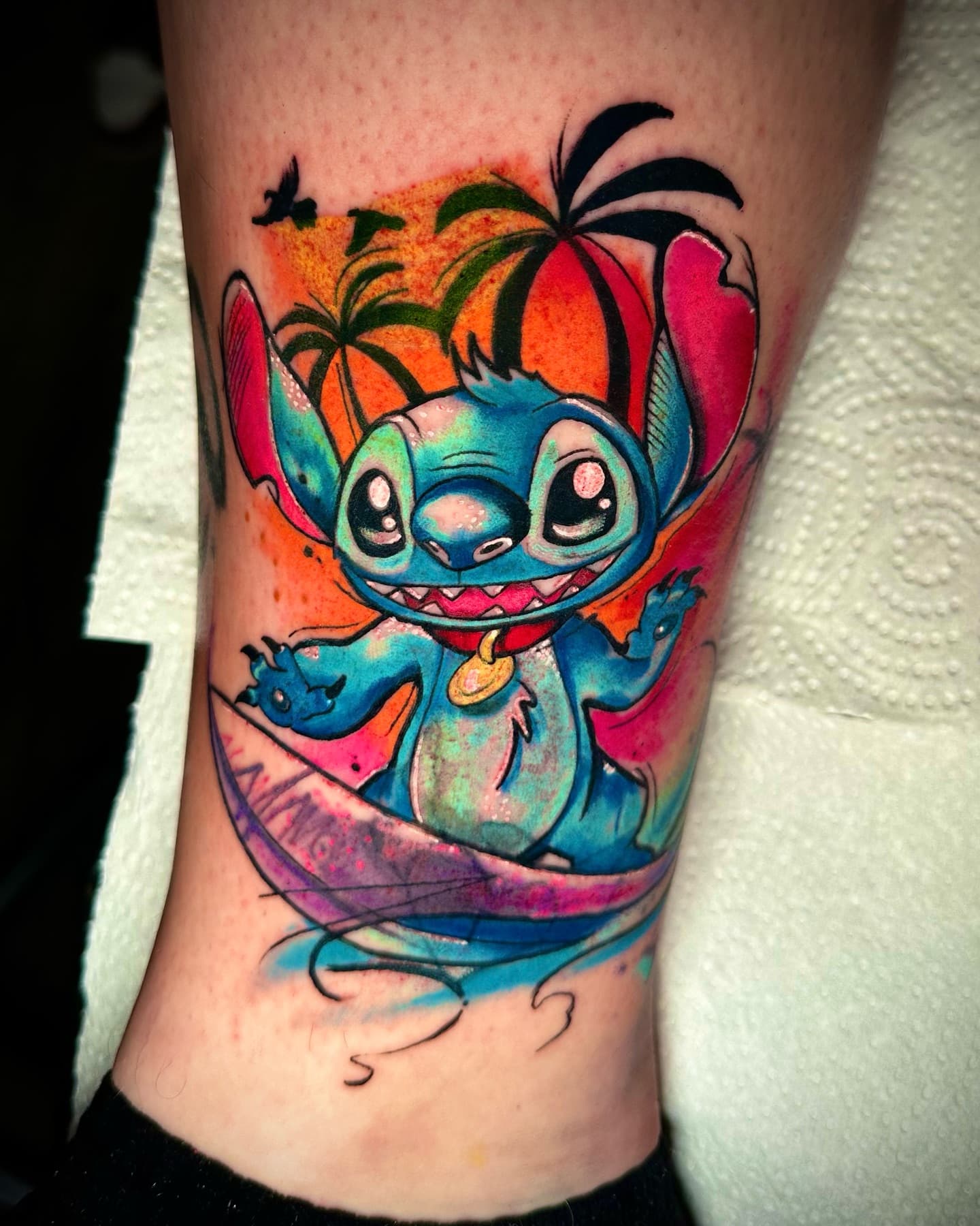 Kleurrijke tattoo van Stitch surfend bij zonsondergang op onderbeen.
