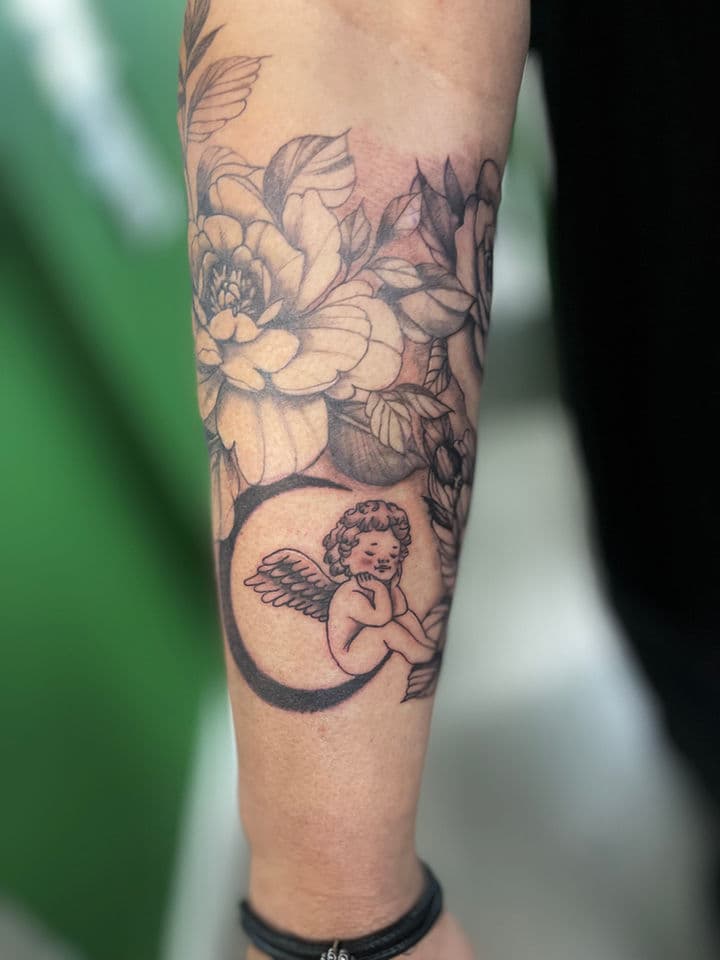 Blackwork tattoo van een cherubijn op een maansikkel met bloemen op de onderarm.