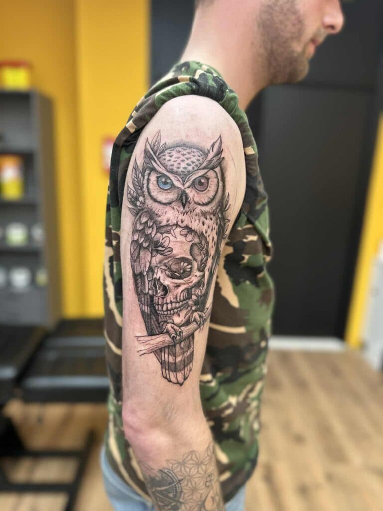 Uil met schedel en bloemen op bovenarm, zwart-wit met gekleurde ogen.