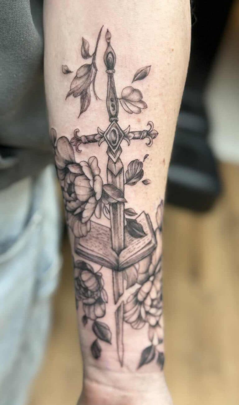 Zwart-wit tattoo van een zwaard door een open boek, omringd door bloemen.