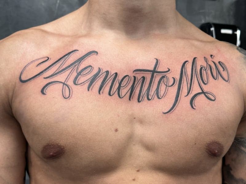 "Memento Mori" tekst tattoo in sierlijk schrift op de borst.
