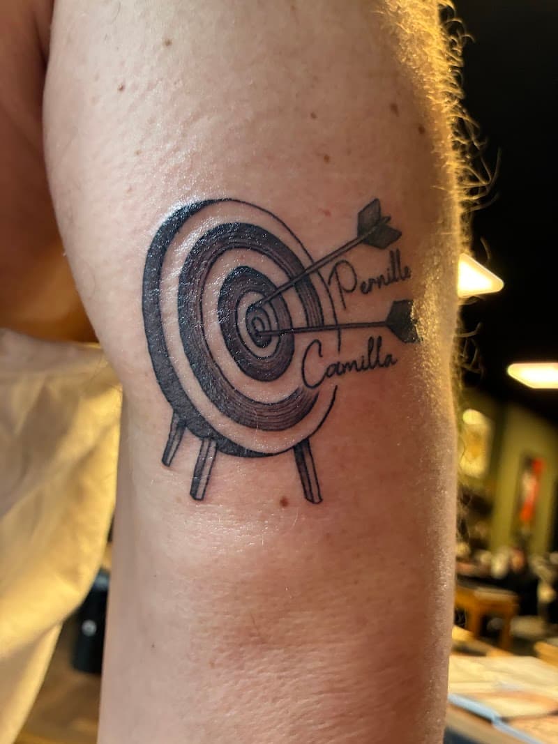 Doelwit tattoo met twee pijlen en namen 'Pernille' en 'Camilla'.