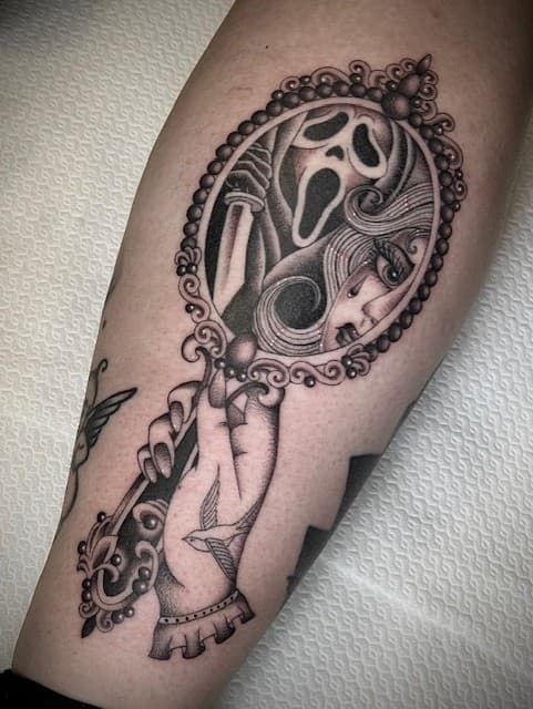 Zwart-wit tattoo van een hand die een spiegel vasthoudt met Ghostface en een huilend meisje.