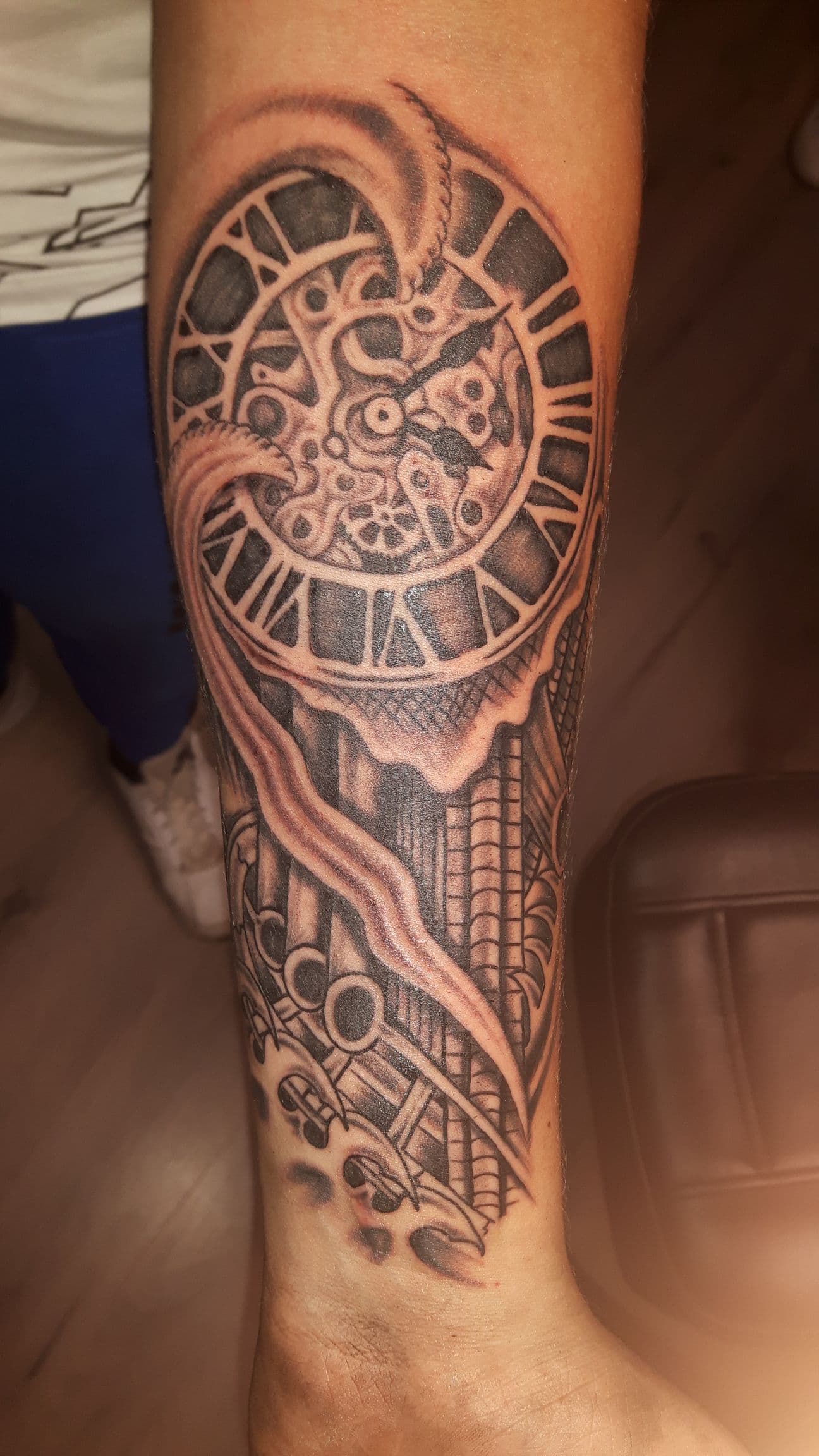 Zwart-wit tattoo van een klok met Romeinse cijfers en tandwielen op de onderarm.