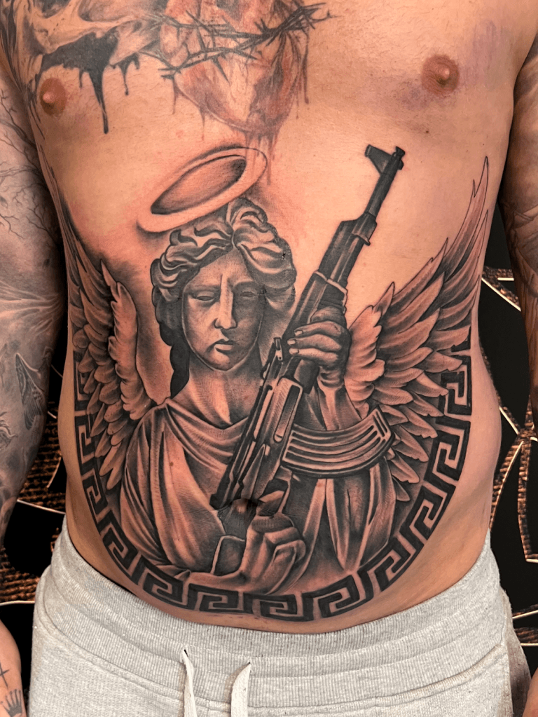 Grote tattoo van een engel met een AK-47 geweer op de buik.