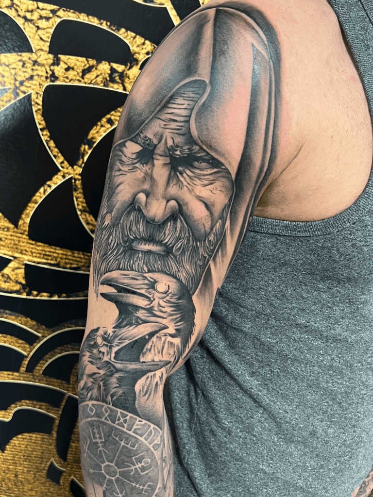 Realistische zwart-wit tattoo van Odin met twee raven en een vegvisir op de bovenarm.