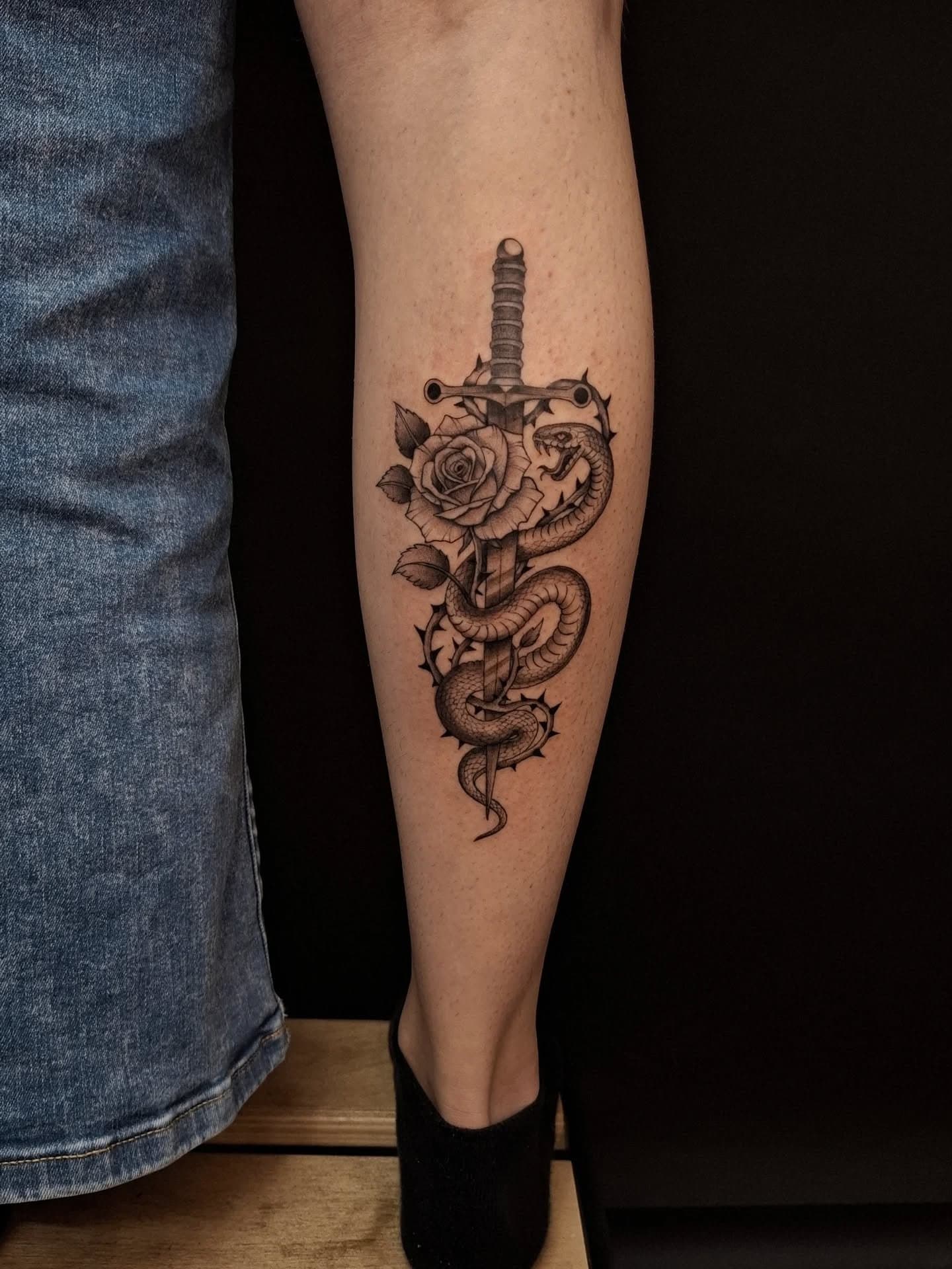 Zwart-wit tattoo van een slang die om een dolk en een roos kronkelt op het onderbeen.