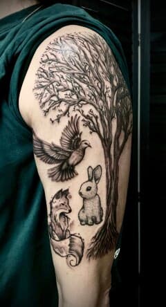 Zwart-wit tattoo van een boom, duif, vos en konijn op de bovenarm.