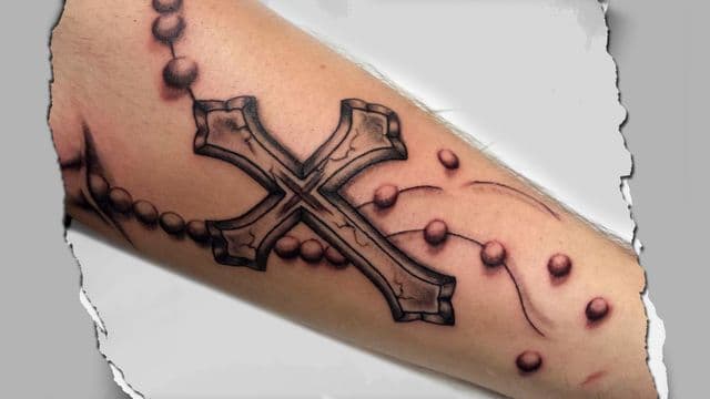 Realistische zwart-grijze tattoo van een kruis met rozenkrans op de arm.