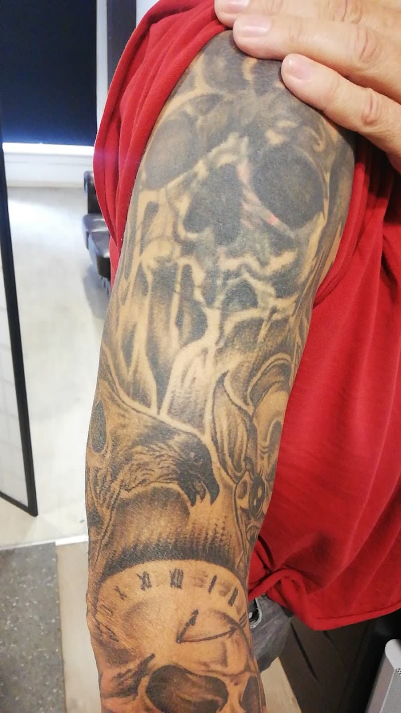 Zwart-wit tattoo van schedel, adelaar en klok op bovenarm.