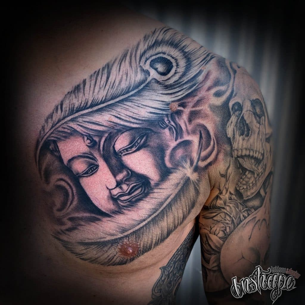 Grote zwart-wit tattoo van Boeddha gezicht met pauwenveer op borst en schouder.