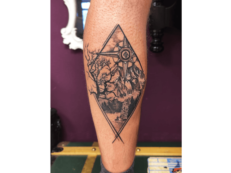 Gedetailleerde kompas en berglandschap tattoo in ruitvorm op onderbeen.