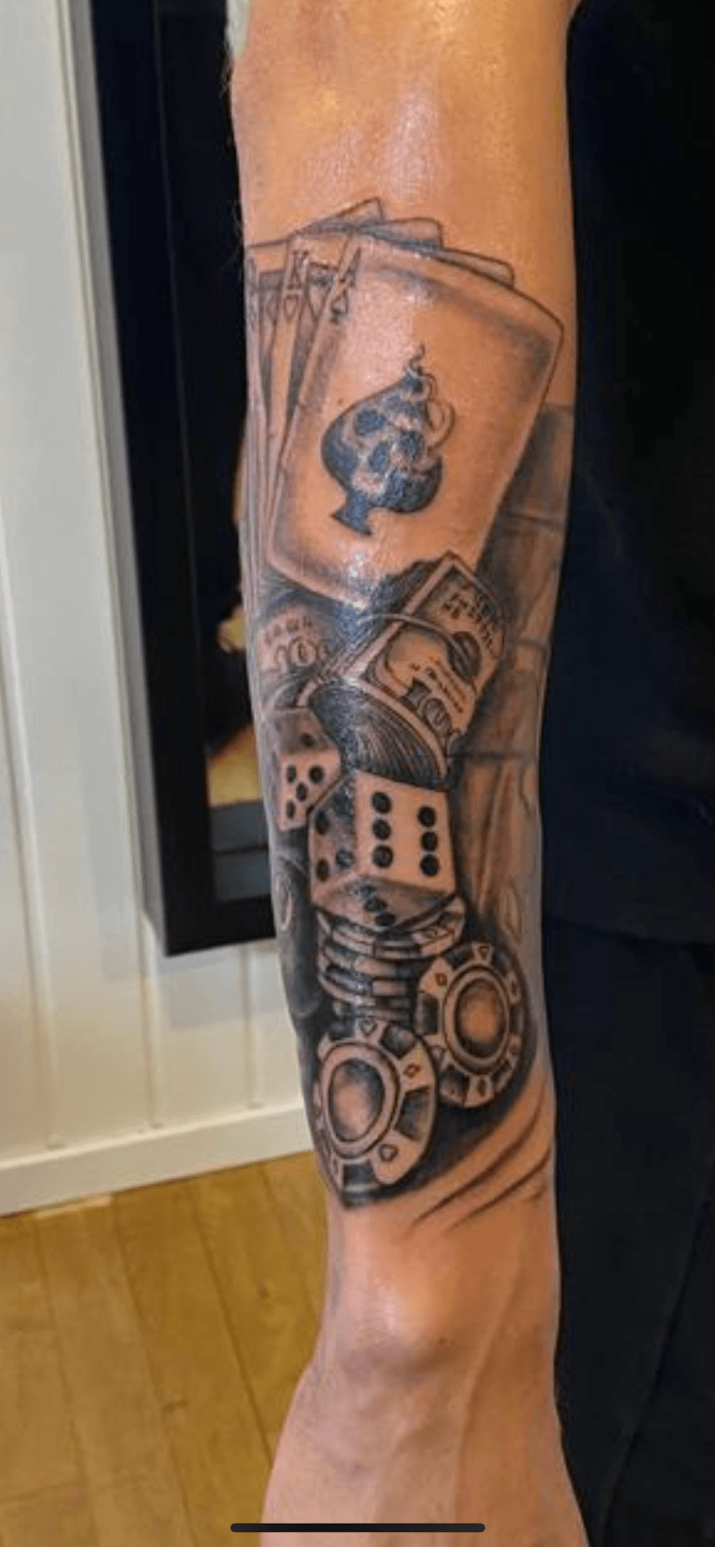 Zwart-wit tattoo van speelkaarten, dobbelstenen, geld en fiches op onderarm.