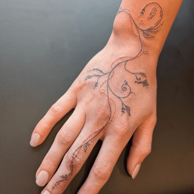Fineline tattoo van sierlijke takken en bladeren op hand en vinger.