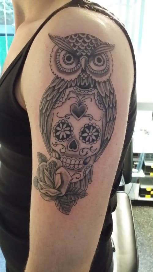 Zwart-wit tattoo van een uil, sugar skull en roos op de bovenarm.