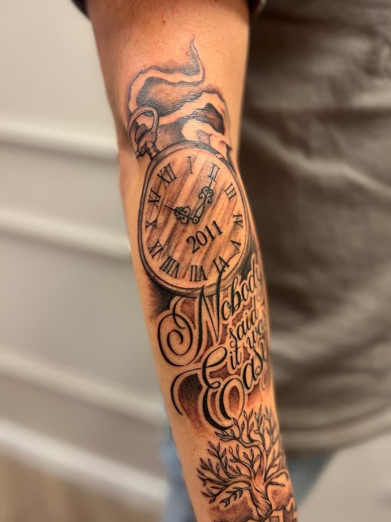 Zwart-wit tattoo van een zakhorloge, tekst en levensboom op de onderarm.