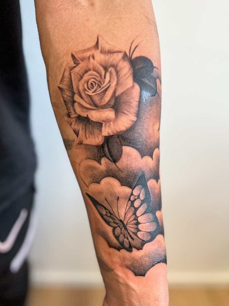 Zwart-wit tattoo van een roos en vlinder met wolken op de onderarm.