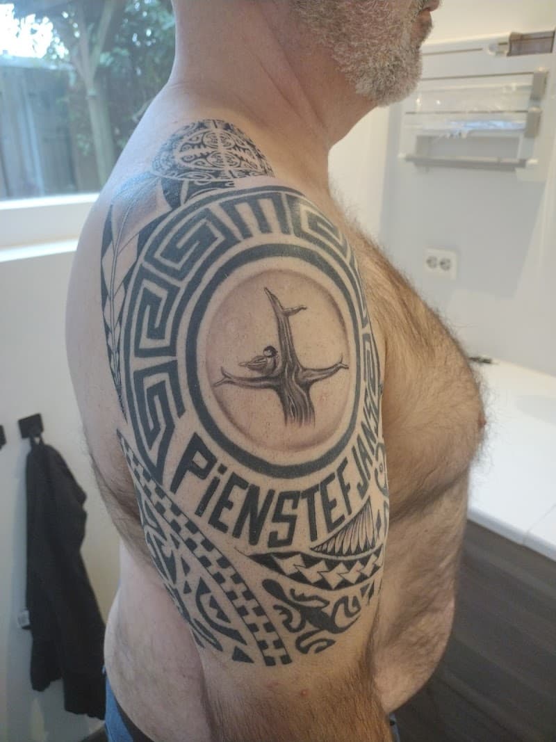 Grote tribal en Griekse tattoo op bovenarm met boom, vogel en namen.