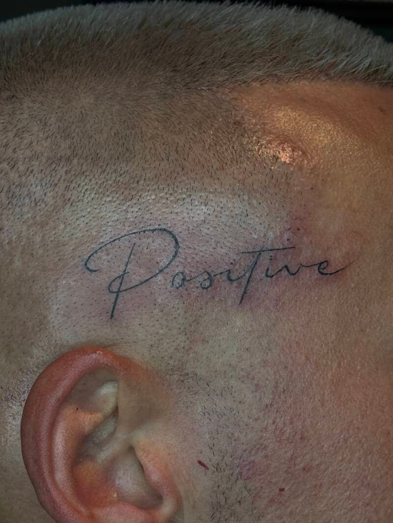 Fineline tekst tattoo 'Positive' op het hoofd boven het oor.