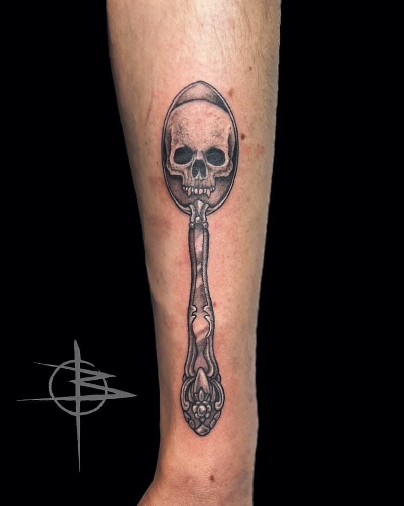 Blackwork tattoo van een schedel in een sierlijke lepel op de onderarm.