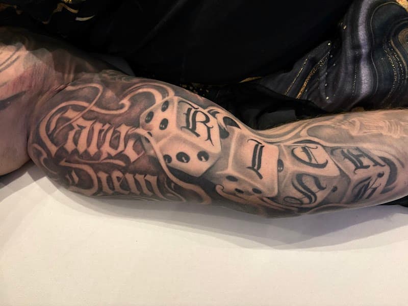 Zwart-wit tattoo op bovenarm met 'Carpe Diem' tekst en dobbelstenen.