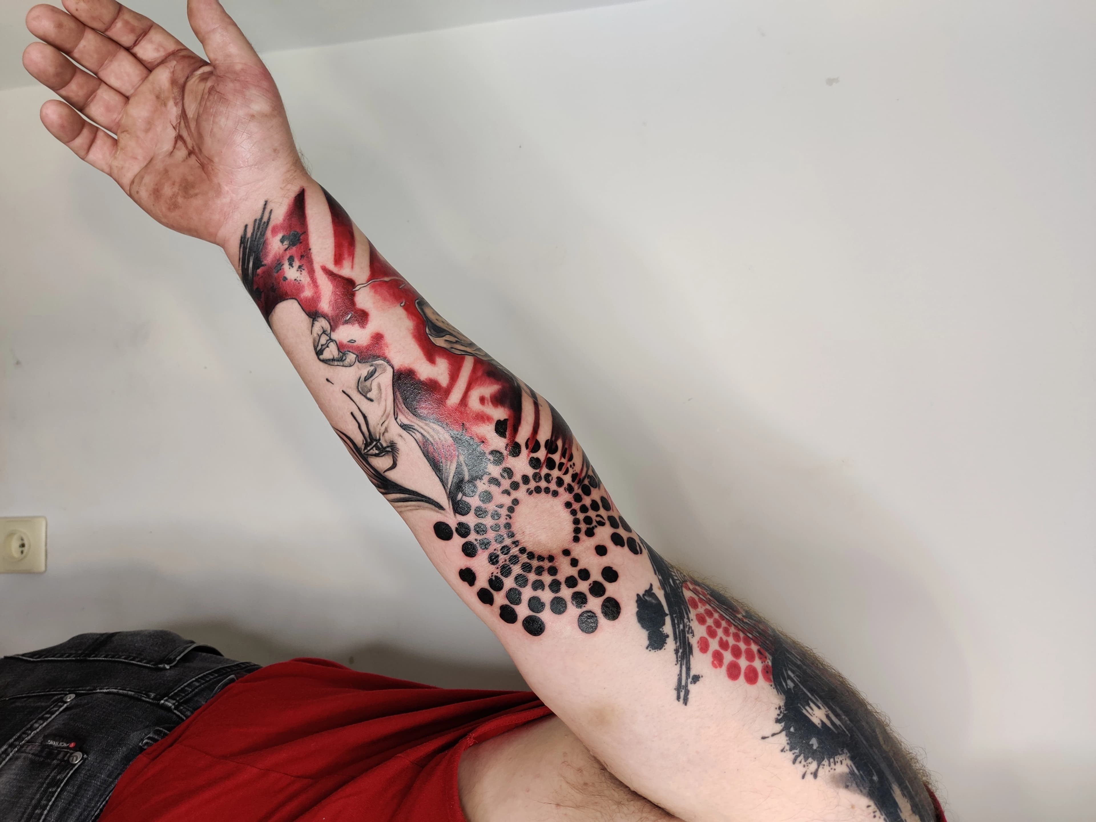 Abstracte tattoo met gezicht, rode en zwarte inkt, en dotwork patroon op onderarm.