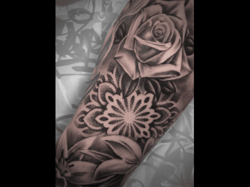 Zwart-wit tattoo van een roos, mandala en bloemen op een bovenarm.