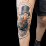 Kleurrijke Dagobert Duck tattoo met hoge hoed en geld op bovenbeen.