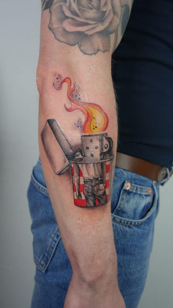 Aansteker tattoo met vlam en persoon in spijkerbroek op de onderarm.