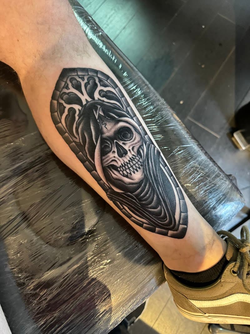 Zwart-wit tattoo van een schedel in een gotisch raamwerk op het onderbeen.