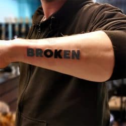 Duidelijke 'BROKEN' tekst tattoo in blokletters op de onderarm.