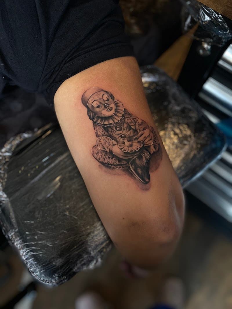 Zwart-wit tattoo van een zittende clown met een bloem op de bovenarm.