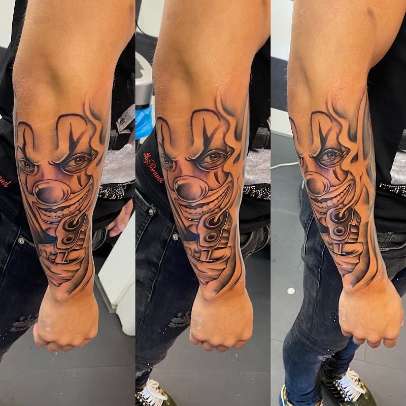 Zwart-wit tattoo van een grijnzende clown met een pistool op de onderarm.